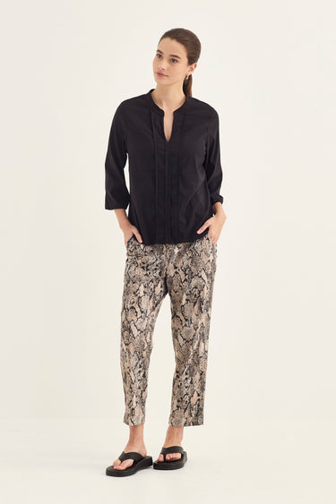 A woman modeling a black long-sleeve button-up shirt and beige-and-brown snakeskin print pants by Verge, paired with black sandals, against a plain off-white studio background.