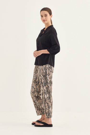 A woman modeling a black three-quarter sleeve top and beige-and-brown snakeskin print wide-leg pants by Verge, paired with black flip-flops, against a plain white studio background.