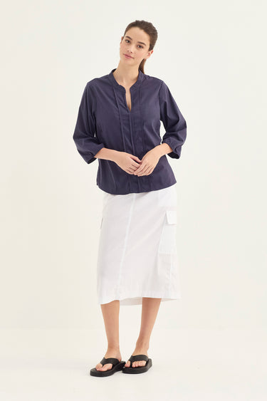 A woman modeling a denim three-quarter sleeve button-up blouse with a split neckline, paired with a white skirt by Verge and black flip-flops, against a plain white studio background.