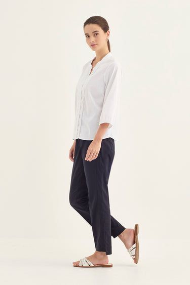 A woman modeling a white button-up shirt and dark navy cropped trousers by Verge, paired with white slide sandals, against a plain off-white studio background.
