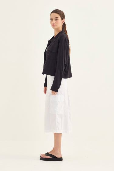 A woman stands in profile wearing a black cropped, long-sleeve jacket, paired with a white midi skirt by Verge and black slide sandals, against a plain off-white studio background.