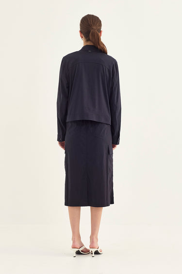 A model viewed from the back wearing a dark navy blue two-piece outfit consisting of a cropped long-sleeve jacket and a matching midi skirt by Verge, paired with white heeled sandals, against a plain off-white studio background.