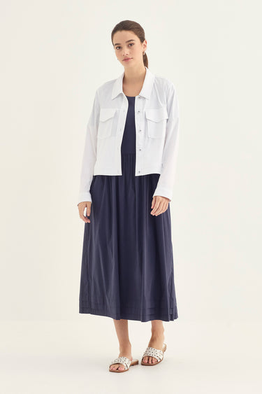 A woman modeling a white cropped button-up shirt jacket over a navy blue midi dress by Verge, paired with silver embellished slide sandals, against a plain off-white studio background.