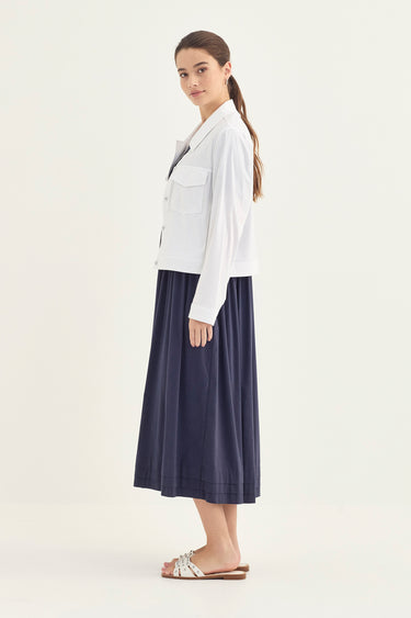A woman modeling a white cropped button-up shirt with chest pockets and a navy midi skirt by Verge, paired with white slide sandals, against a plain off-white studio background.