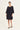 A woman modeling a loose-fitting black shift dress by Verge with elbow-length sleeves and a wide scoop neckline, paired with black sandals, against a plain light gray studio background.