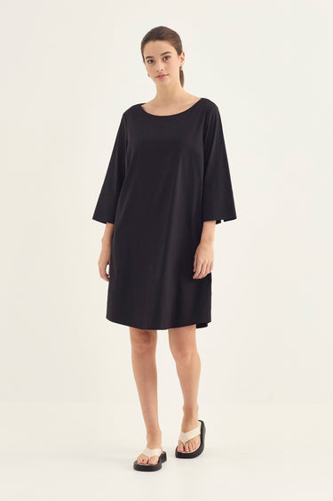 A woman modeling a loose-fitting black shift dress by Verge with elbow-length sleeves and a wide scoop neckline, paired with black sandals, against a plain light gray studio background.