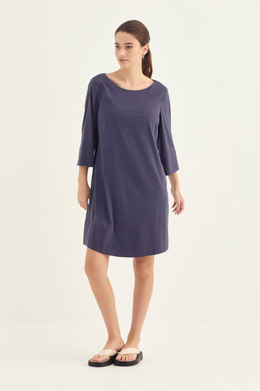 A woman modeling a loose, knee-length dress in a muted periwinkle or dusty purple shade. The dress by Verge has three-quarter sleeves and a relaxed silhouette, paired with black slide sandals, set against a plain off-white studio background.