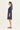 A woman in profile wearing a loose, short-sleeve navy blue tunic dress by Verge and beige slide sandals, standing against a plain off-white studio background.