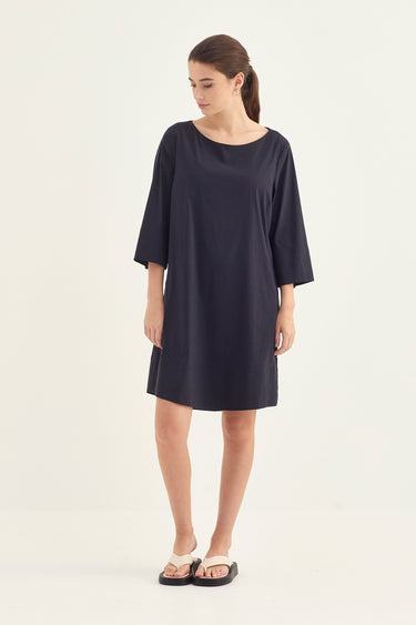 A woman modeling a loose, short-sleeve navy blue dress by Verge with a wide neckline and straight silhouette, paired with black slide sandals, against a plain off-white studio background.