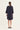 A woman viewed from the back wearing a dark navy blue shift dress by Verge with three-quarter sleeves and black flat shoes, set against a plain off-white studio background.