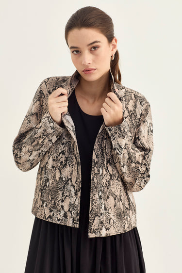 A woman modeling a cropped, long-sleeve jacket with a beige and black snakeskin pattern, worn over a black top and black skirt by Verge against a light gray studio background.