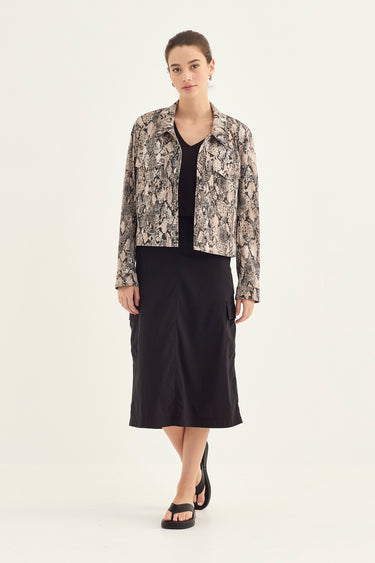 A model stands against a plain light background wearing a black midi skirt, a black top, and a cropped long-sleeve jacket by Verge with a beige and black snakeskin pattern.
