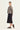 A model is shown in profile wearing a cropped, long-sleeve jacket with a beige, black, and gray snake print, paired with a black midi skirt by Verge and black flip-flop sandals, against a plain light background.