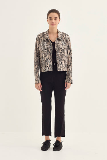 A woman stands against a plain light background wearing a cropped long-sleeve button-up shirt with a beige, black, and gray animal print, a black top underneath, black cropped pants by Verge, and black shoes.