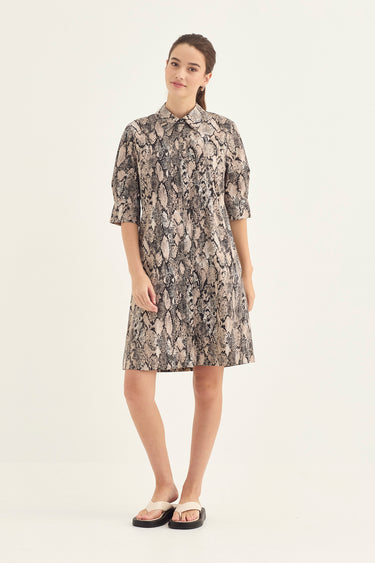 A woman modeling a short-sleeve collared dress by Verge with an all-over abstract print in beige, gray, and brown tones, paired with black slide sandals, against a plain light background.