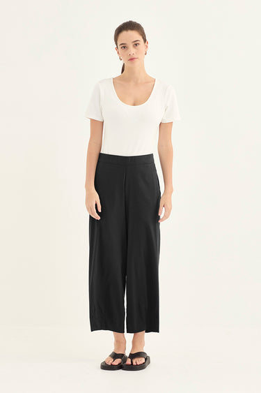 A woman stands against a plain white studio background wearing a white short-sleeve scoop-neck top, black wide-leg cropped pants by Verge, and black sandals.