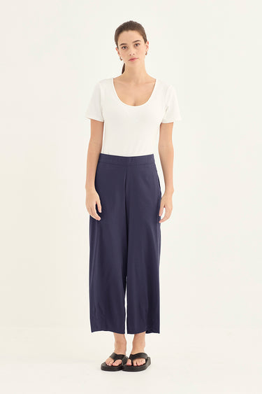 A woman modeling a white short-sleeve scoop-neck top and navy blue wide-leg cropped pants by Verge, paired with black sandals, against a plain white studio background.