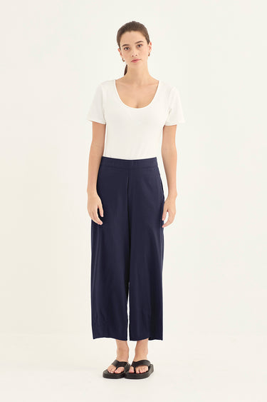 A woman stands against a plain light background wearing a white short-sleeve scoop-neck top, navy wide-leg cropped pants by Verge, and black sandals.
