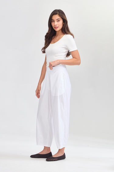 A woman modeling a white short-sleeve top and white wide-leg cropped pants by Verge, paired with dark flat shoes, against a plain light gray studio background.