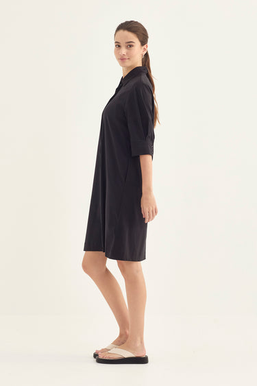 A woman modeling a black short-sleeve collared shirt dress by Verge with side pockets, paired with beige slide sandals, against a plain off-white studio background.