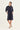 A woman modeling a dark navy blue shirt dress with three-quarter sleeves by Verge, a pointed collar, and side pockets, paired with beige flat sandals against a plain off-white studio background.