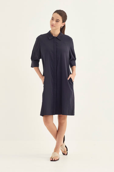 A woman modeling a dark navy blue shirt dress with three-quarter sleeves by Verge, a pointed collar, and side pockets, paired with beige flat sandals against a plain off-white studio background.