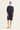 A woman modeling a dark navy blue oversized shirt dress by Verge with three-quarter sleeves and a collar, paired with beige flat sandals, against a plain off-white studio background.