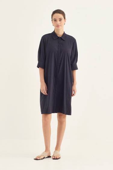 A woman modeling a dark navy blue oversized shirt dress by Verge with three-quarter sleeves and a collar, paired with beige flat sandals, against a plain off-white studio background.