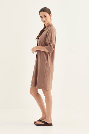 A woman modeling a brown short-sleeve shirt dress by Verge with a relaxed fit, standing against a plain off-white studio background.