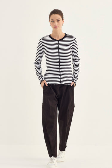 A woman modeling a black and white horizontally striped zip-front cardigan with black trim, paired with black pants by Verge and white sneakers, against a plain light background.
