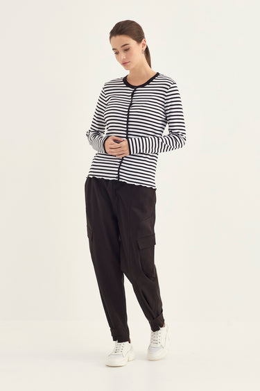 A woman modeling a black and white horizontally striped zip-up long-sleeve top, paired with black cargo pants by Verge and white sneakers, against a plain off-white studio background.