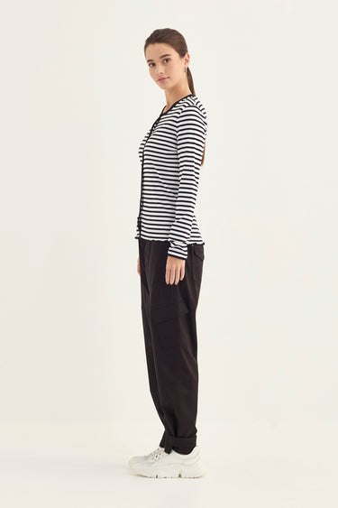 A woman modeling a black and white horizontally striped long-sleeve top with a crew neck, paired with black pants by Verge and white sneakers, standing against a plain off-white studio background.