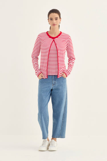 A woman modeling a red and white striped long-sleeve cardigan with red trim, paired with light blue jeans by Verge and white sneakers, against a plain off-white studio background.