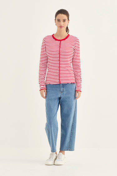 A woman stands against a plain light background wearing a red and white horizontally striped zip-front cardigan by Verge with red trim, light blue jeans, and white sneakers.