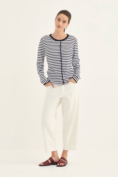 A woman modeling a black and white horizontally striped long-sleeve top with black trim, paired with white cropped pants by Verge and dark brown slide sandals, against a plain white studio background.