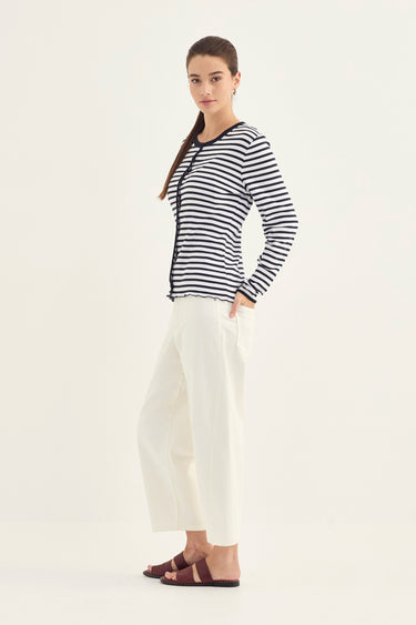 A woman modeling a black and white horizontally striped long-sleeve top with a round neckline, paired with white cropped wide-leg pants by Verge and dark brown slide sandals, set against a plain off-white studio background.