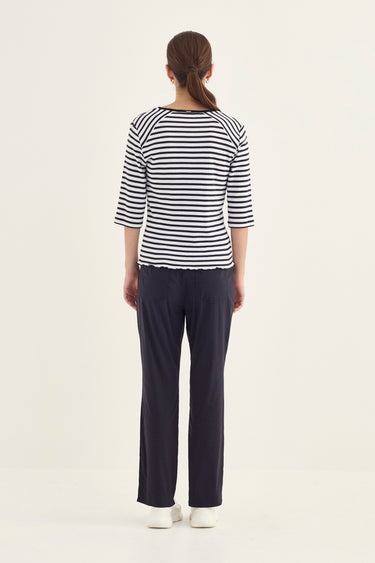 A woman viewed from the back wearing a navy and white horizontally striped 3/4-sleeve top, dark navy pants by Verge, and white shoes, against a plain off-white studio background.