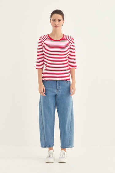 A woman modeling a red and white horizontally striped short-sleeve top with red trim by Verge, paired with light blue wide-leg jeans and white sneakers, against a plain off-white studio background.