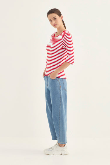 A woman modeling a red and white striped short-sleeve t-shirt, paired with light blue jeans by Verge and white sneakers, standing against a plain white studio background.