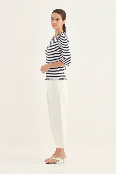 A woman modeling a navy and white horizontally striped short-sleeve top with a scalloped edge, paired with white pants by Verge and white heeled sandals, against a plain white studio background.