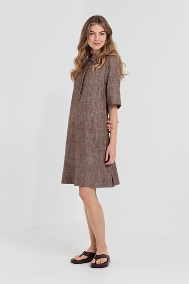 A woman modeling a brown textured linen dress with short sleeves by Verge, a high collar, and a tie-neck detail, paired with dark brown slide sandals, against a plain light gray studio background.