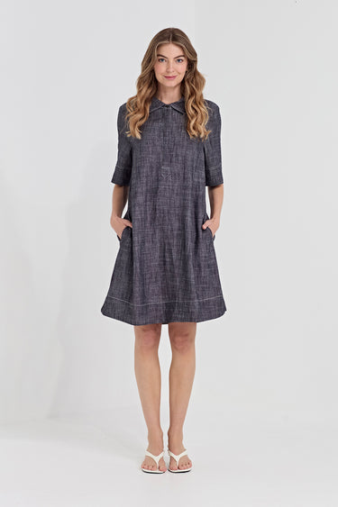 A woman modeling a dark gray textured linen dress by Verge with a collar, 3/4 sleeves, and side pockets, standing against a light gray studio background.