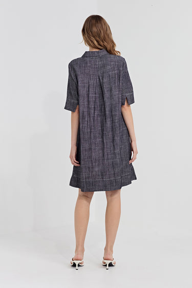 A woman viewed from the back wearing a short-sleeve plaid shirt dress by Verge in dark gray and black, paired with white heeled sandals, against a light gray studio background.