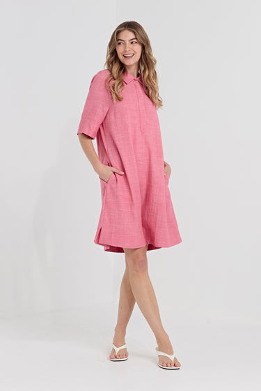 A woman modeling a pink short-sleeve linen dress with a high collar and side pockets by Verge, paired with white thong sandals, against a light gray studio background.