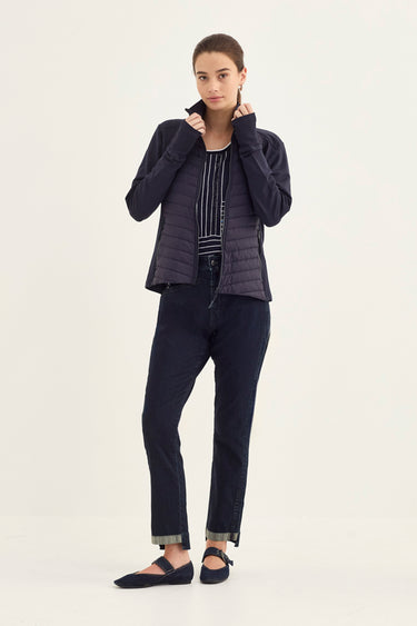 A woman modeling a dark navy blue outfit consisting of a zippered puffer jacket, a striped top, slim-fit jeans by Verge, and flat shoes, photographed against a solid off-white studio background.