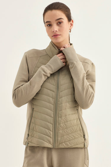A woman wearing a light beige, quilted zip-up jacket with integrated sleeves and matching pants by Verge, posed against a solid off-white studio background.