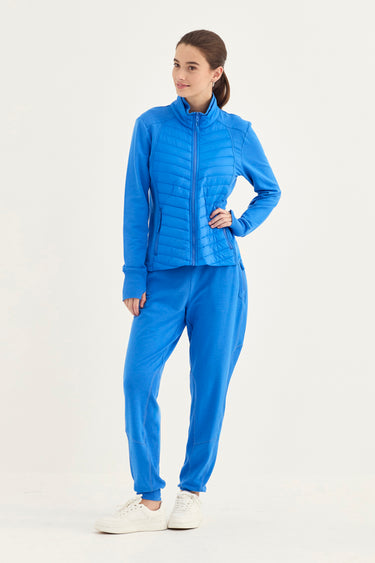 A woman modeling a matching bright blue tracksuit consisting of a zip-up puffer jacket and jogger pants, paired with white sneakers by Verge, against a plain white studio background.