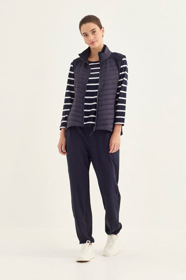 A woman modeling a dark navy blue puffer vest over a navy and white striped long-sleeve shirt, paired with navy jogger pants by Vergeand white sneakers, against a plain light gray studio background.