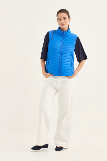 A model stands against a plain white studio background wearing a bright blue quilted zip-front puffer vest over a black short-sleeve top, paired with white wide-leg pants and black flats by Verge.