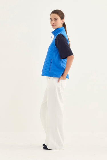 A woman modeling a bright blue quilted puffer vest over a black short-sleeve top and white wide-leg pants by Verge, standing against a plain white studio background.